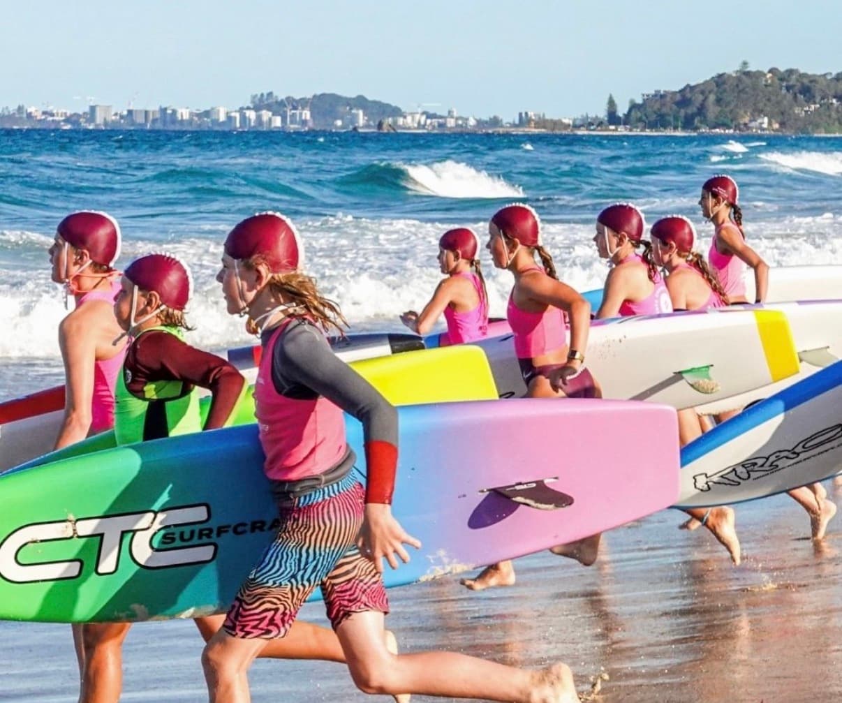 Nippers in maroon caps running into the surf carrying racing boards